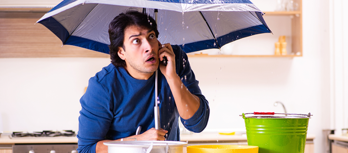 A worried man holding blue umbrella as his ceiling leaks into multiple pots sitting on the counter of his kitchen.